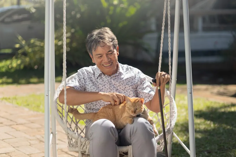 An elderly man holding a ginger cat on his lap while sitting on a swing.