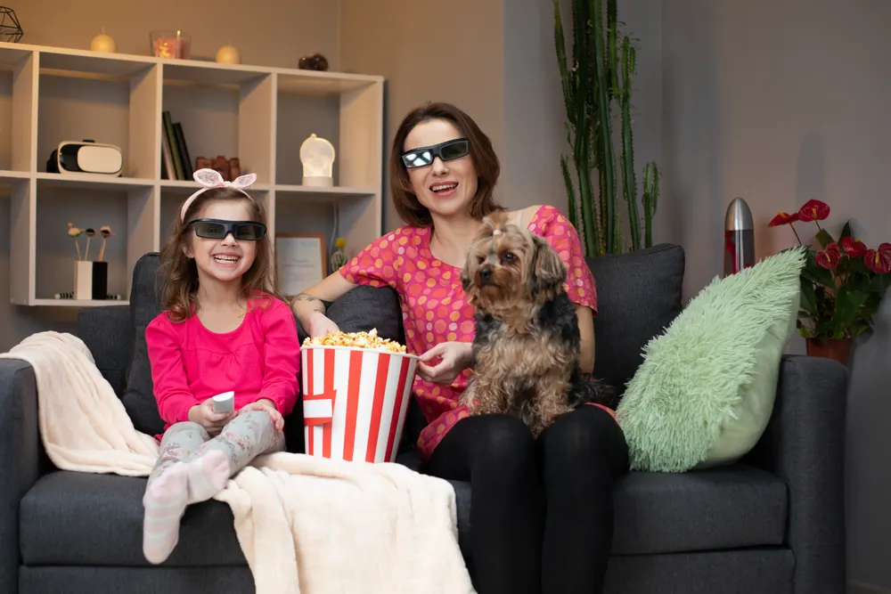 A mum with her dog on her lap and her daughter next to her in 3D glasses watching a film while eating popcorn.